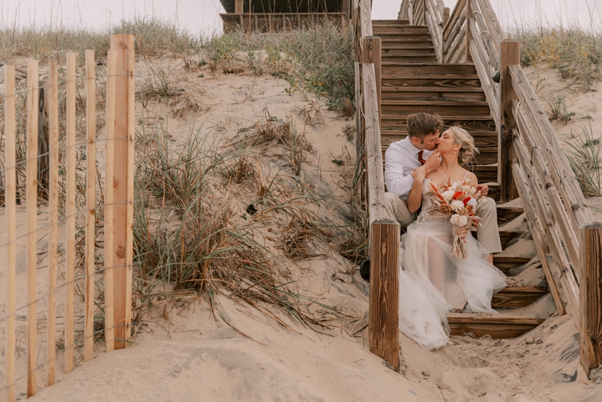 bride sits between groom's knees on wooden beach steps