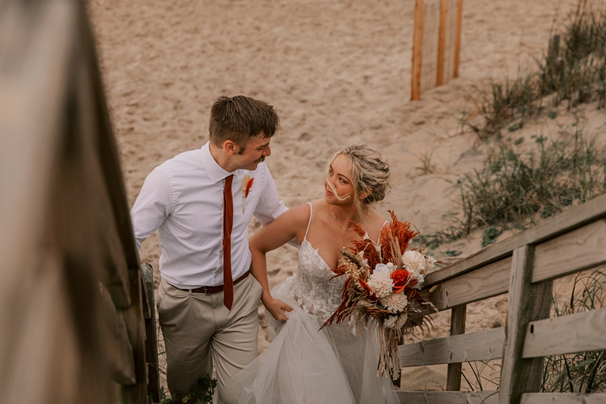 groom holds bride's hand walking up wooden steps on Kill Devil Hills beach