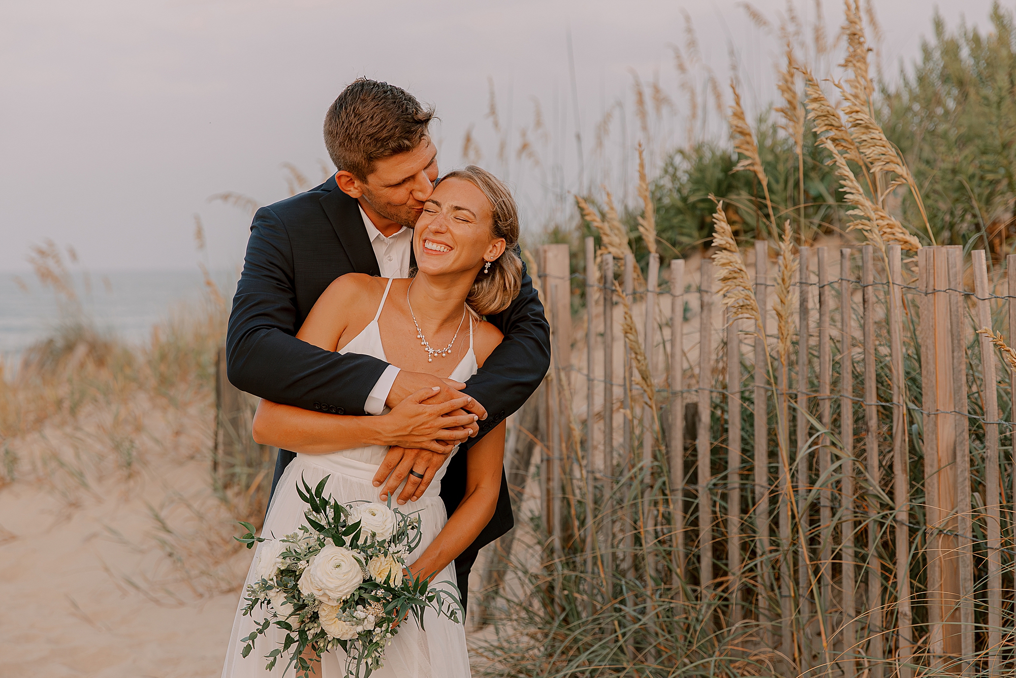 groom hugs bride from behind making her laugh between sand dunes 