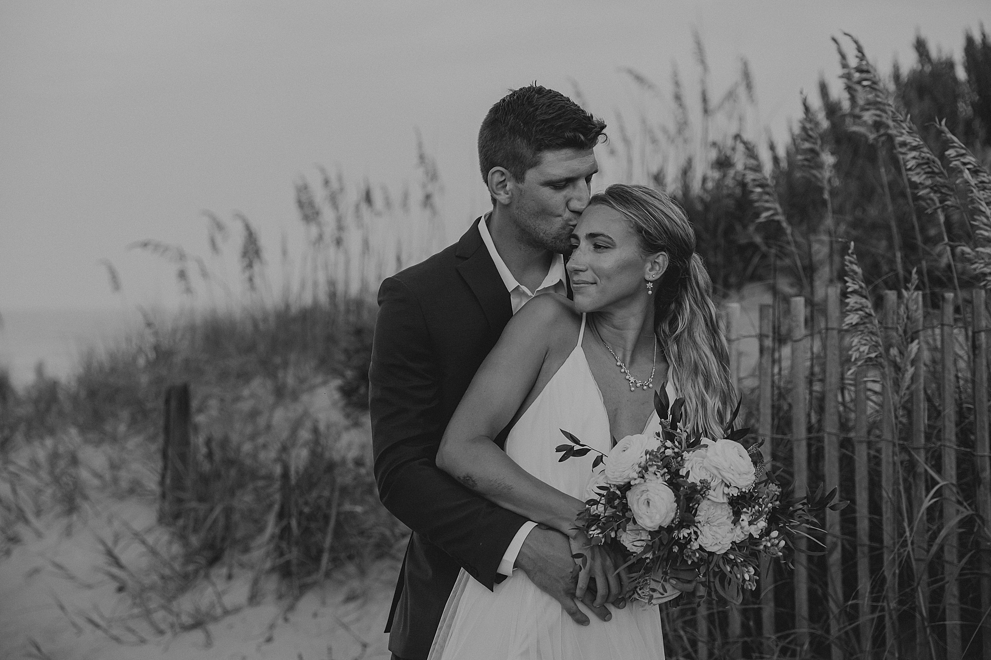groom hugs bride from behind in front of sand dunes in kill devil hills