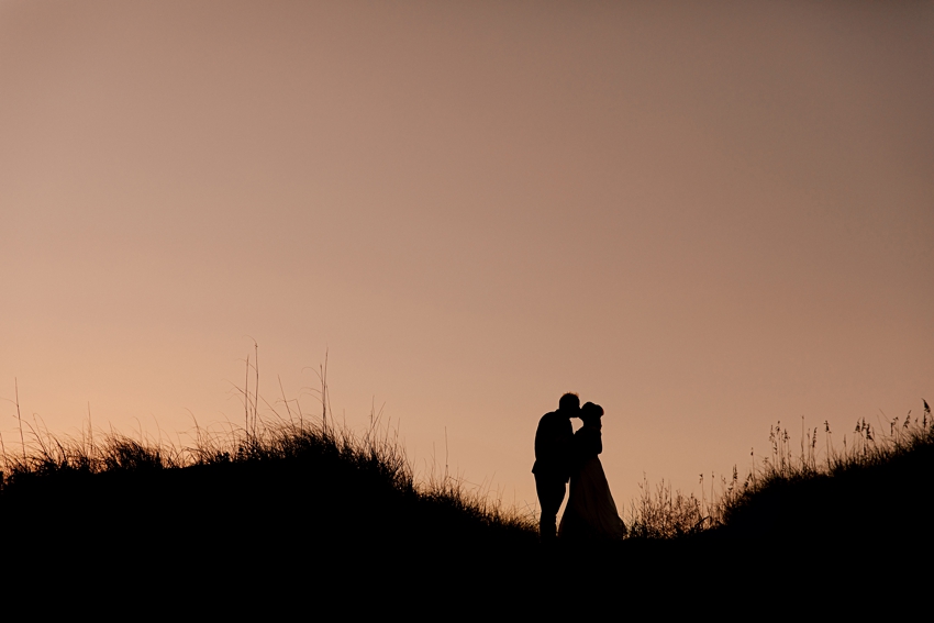 silhouette of bride and groom on sand dunes with beach grass with peach sunset