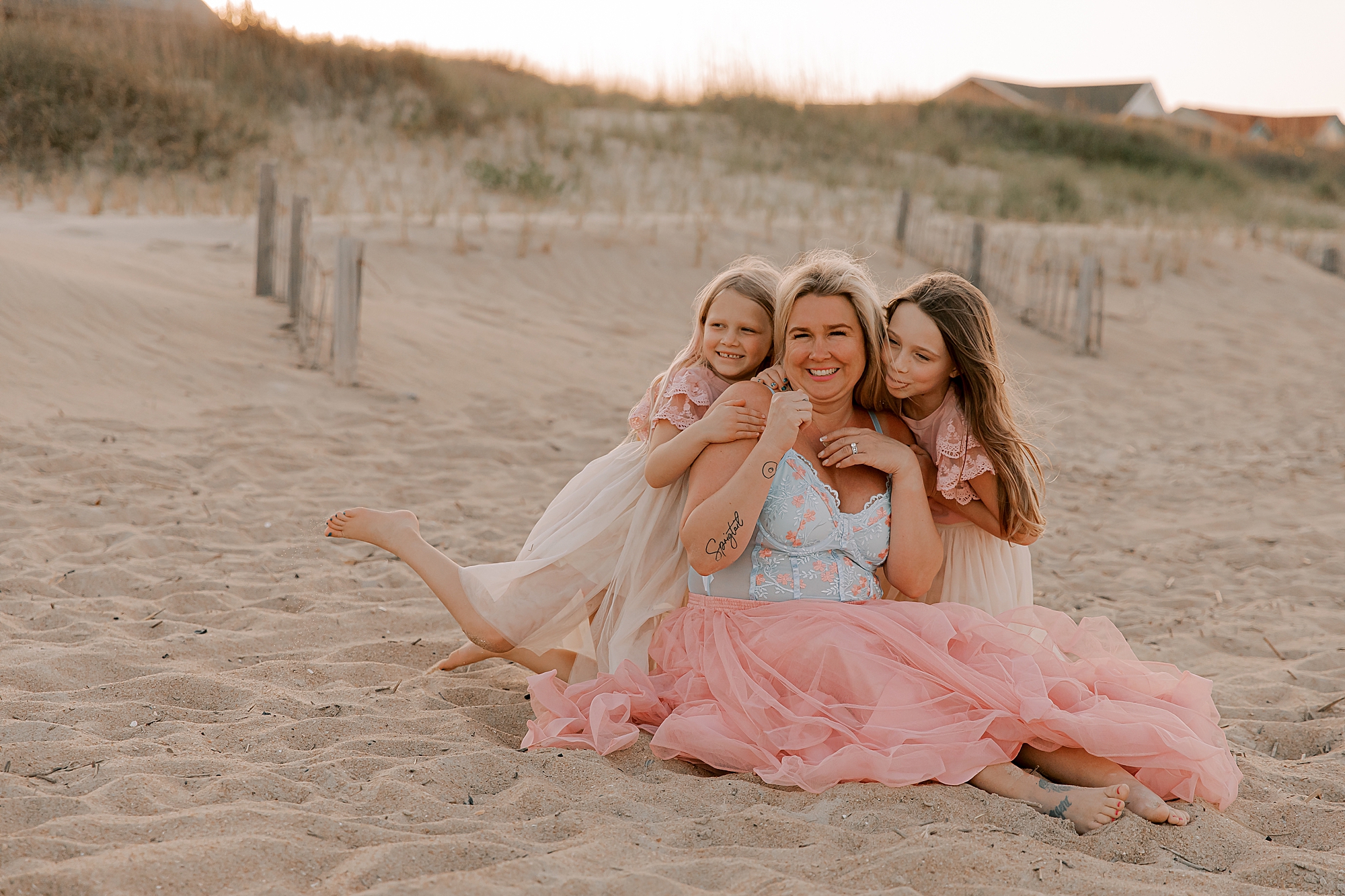 woman in pink skirt hugs two daughters in matching dresses during mommy + me photos at nags head pier