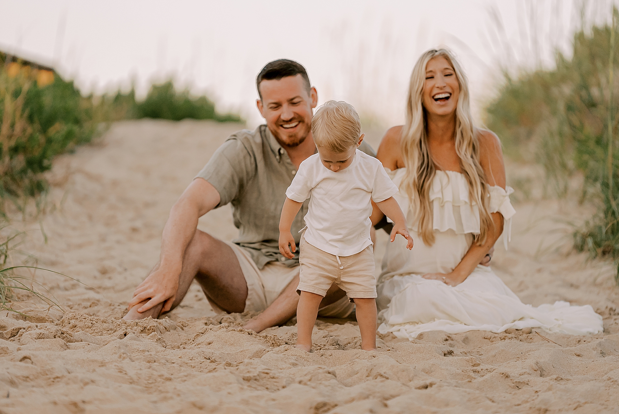 parents laugh while toddler stand in front of them on the sand