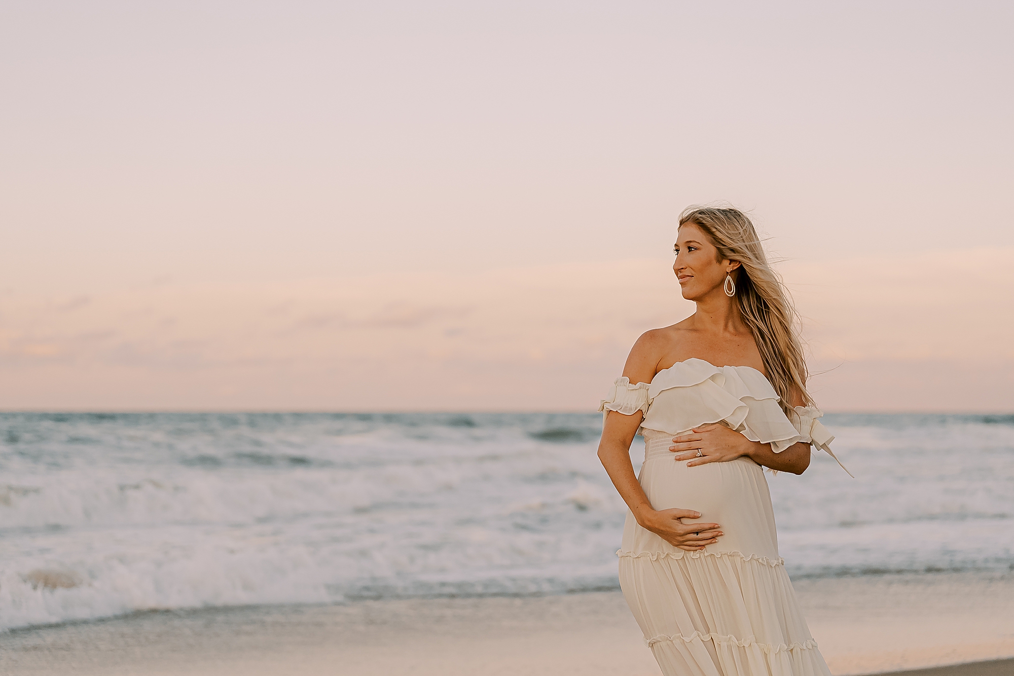 mom in white dress holds baby bump at sunset on the beach