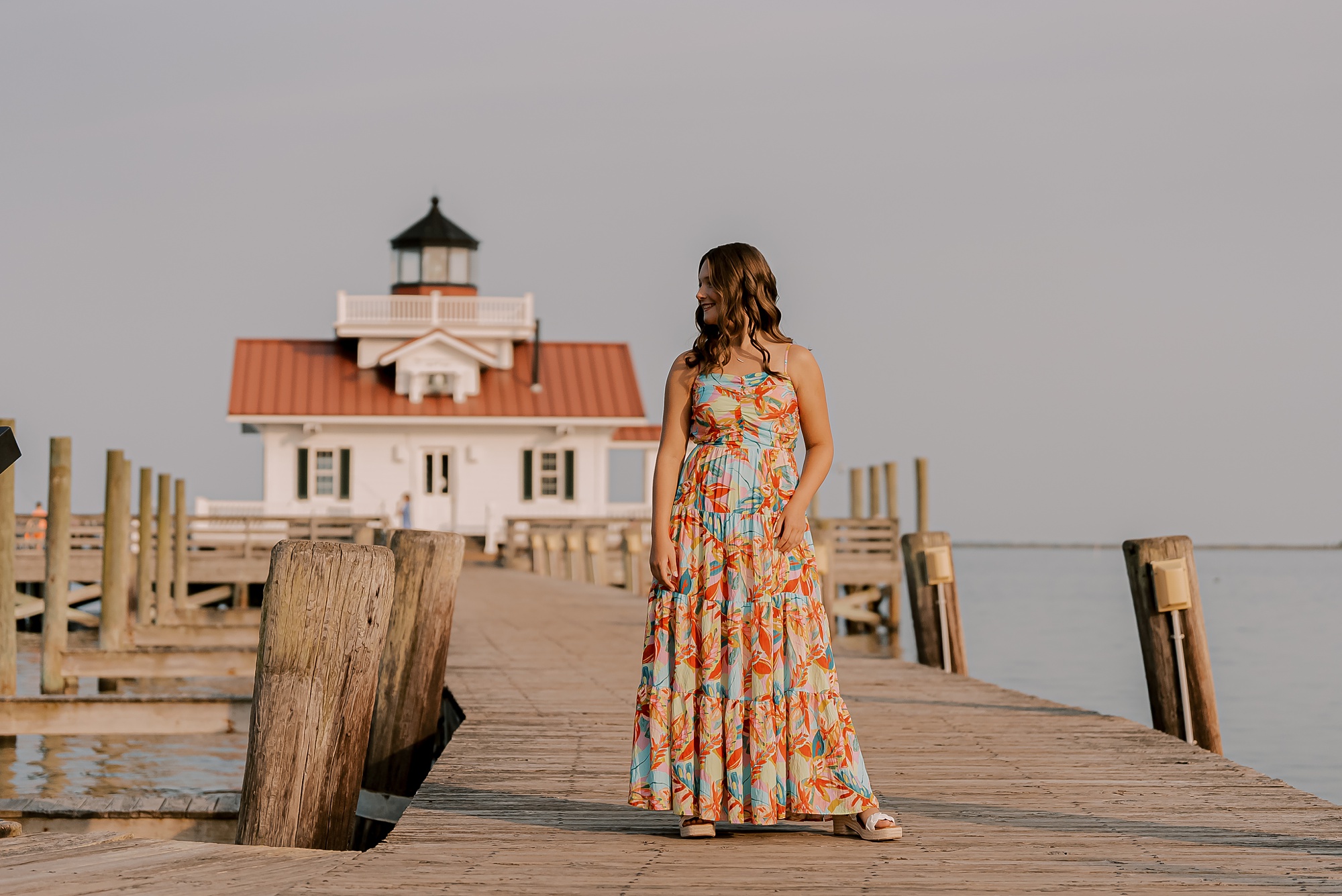 young woman stands on wooden dock in front of Manteo lighthouse