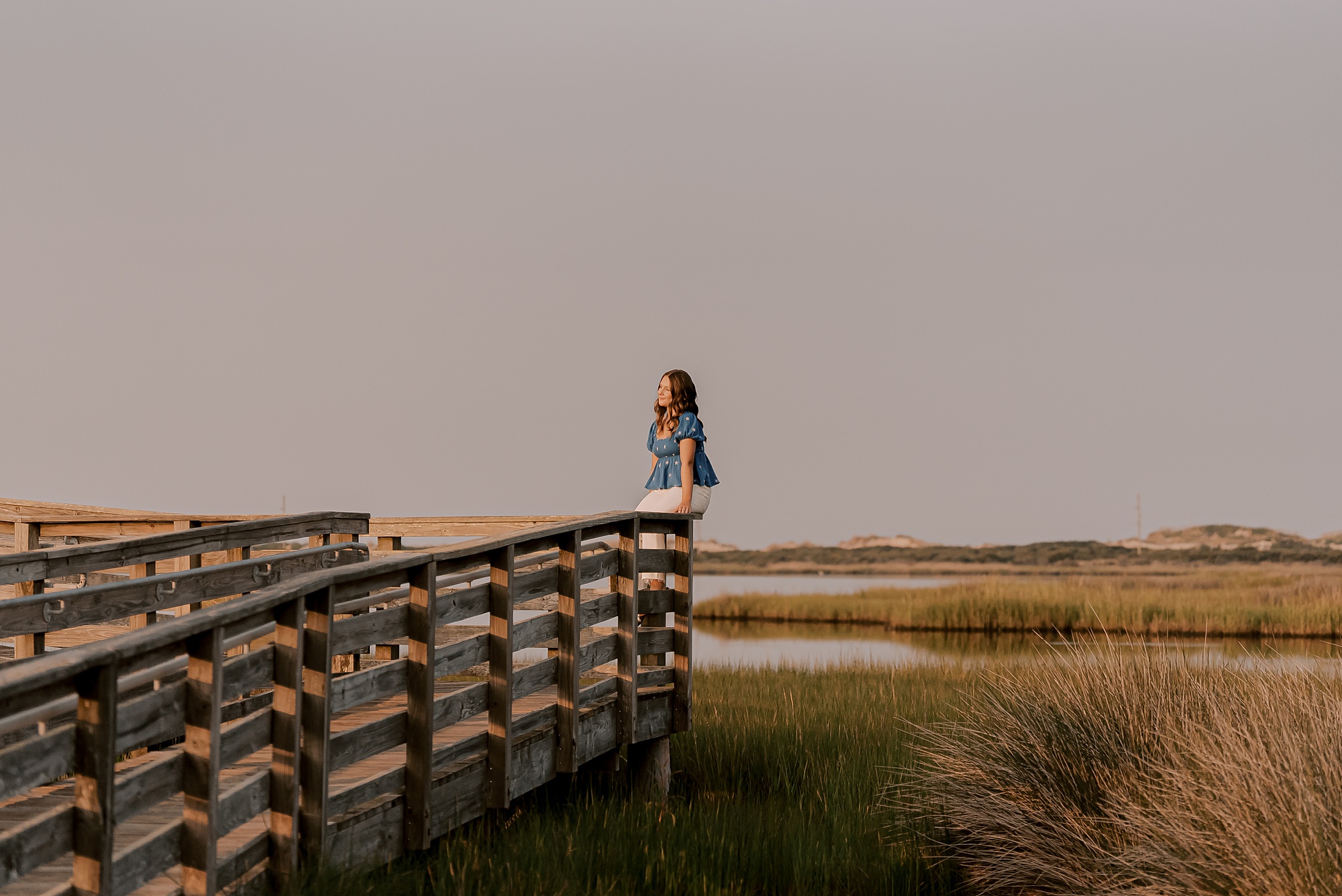 girl sits on wooden dock in marsh during outer banks senior portraits