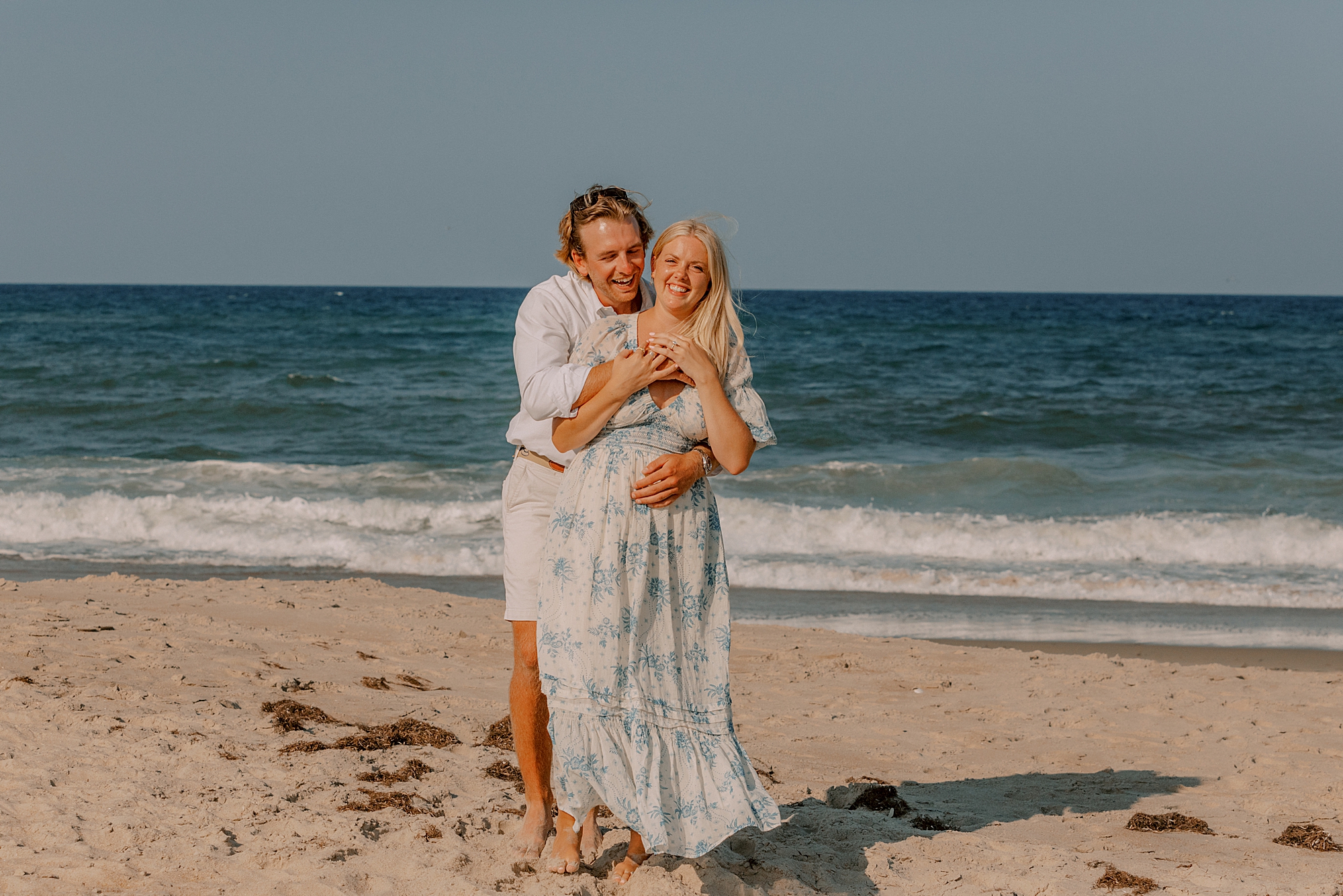 man in white shirt hugs woman from behind in front of ocean