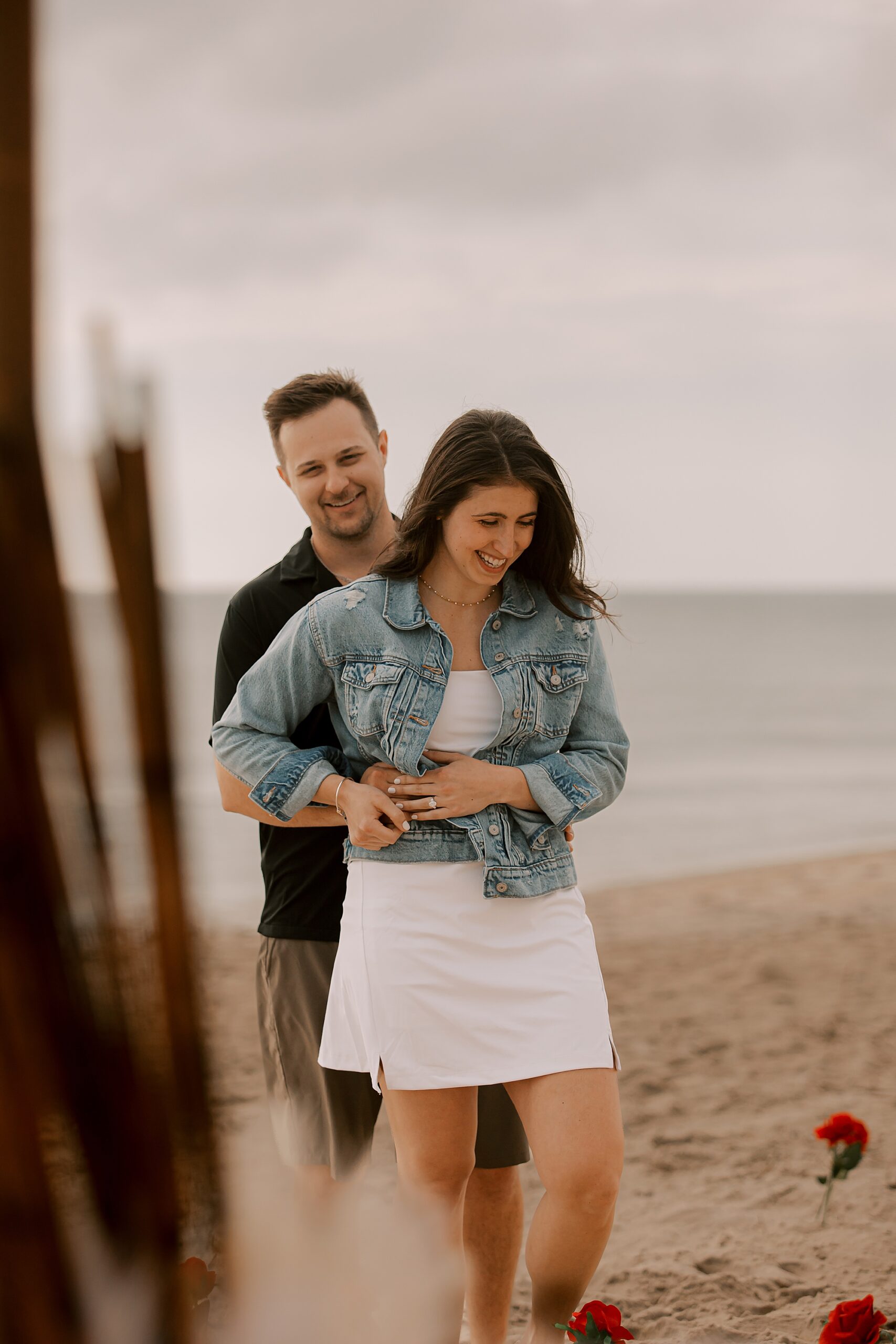 man hugs woman in white dress with jean jacket from behind on kitty hawk beach