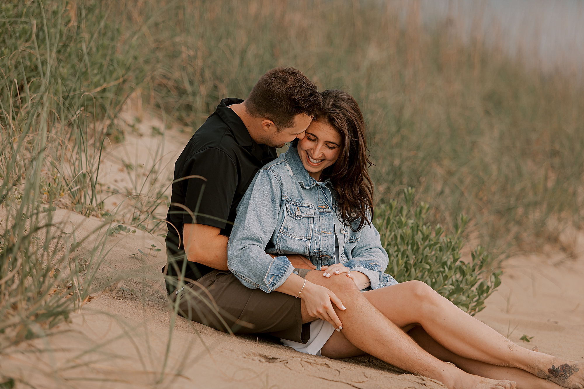 engaged couple sits on sand dune on kitty hawk beach