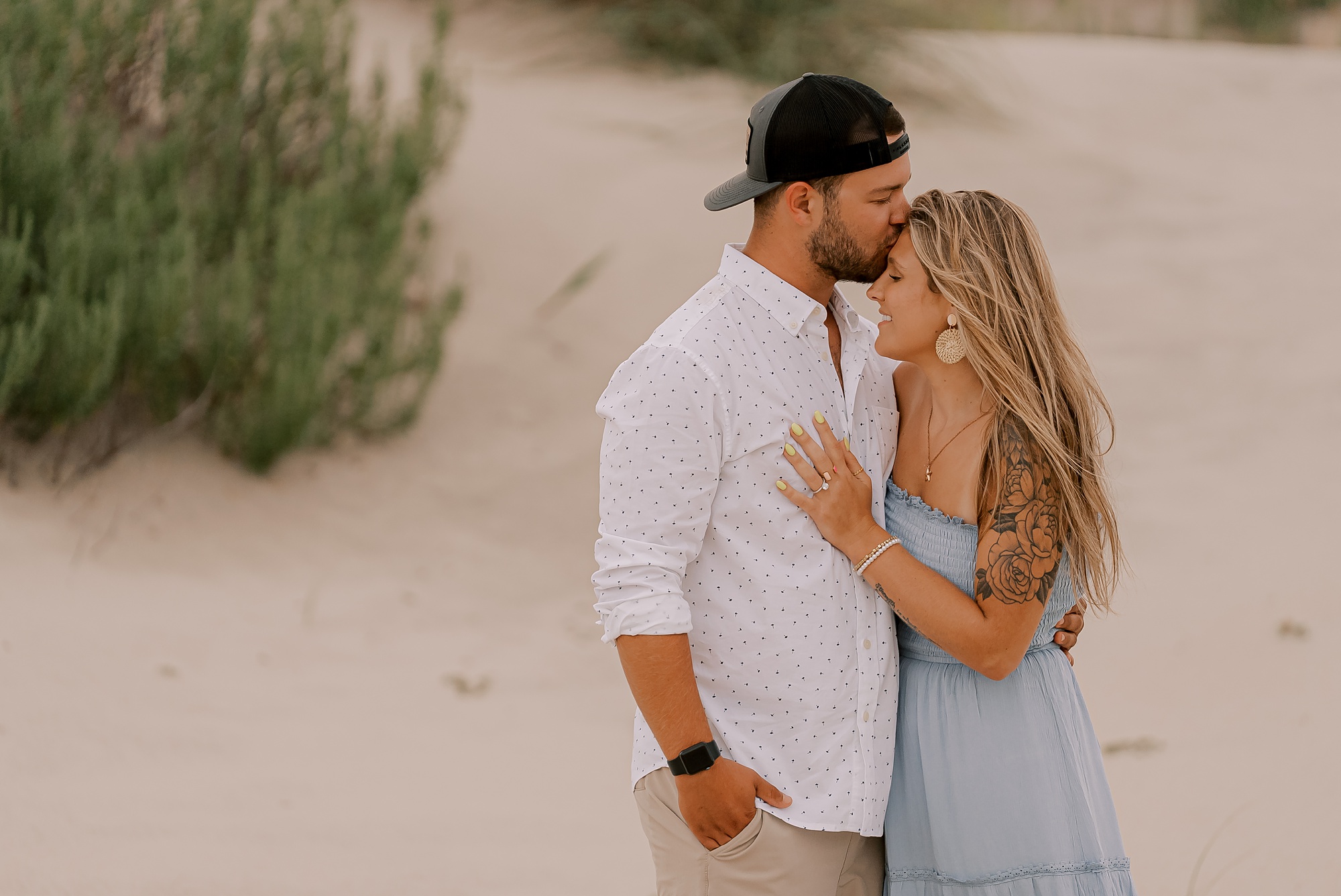 man hugs fiancee to him kissing her forehead on the beach